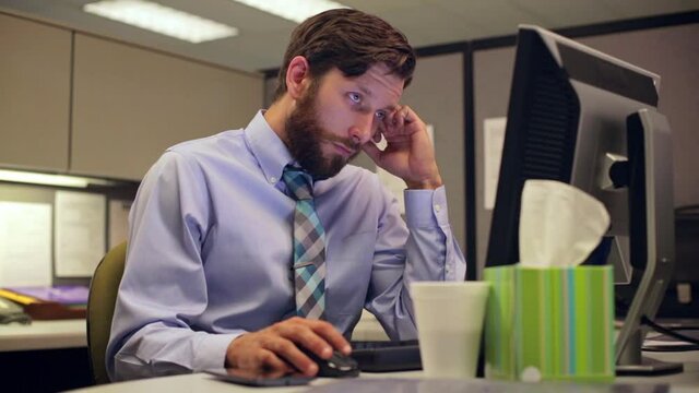 A sick young Caucasian man working in his cubicle office, fighting off a cold with tissues and hot tea at his computer.
