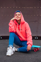 Portrait of a trendy pretty young girl sitting next to the skateboard coart on her plastic...
