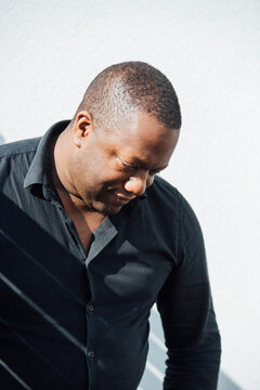 A black man in his late 30s, standing in the sun, looking down.