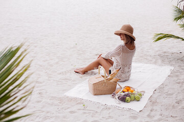 A young woman sits on the beach carpet in a straw hat and a white knitted clothes with back to the camera
 