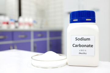 Selective focus of a bottle of sodium carbonate chemical compound or soda ash beside a petri dish with solid crystalline powder substance. White Chemistry laboratory background with copy space. - obrazy, fototapety, plakaty