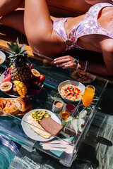 Fit pretty woman with perfect tanned bronze body skin lies on edge of pool  with floating breakfast on vacation in Thailand. 