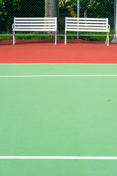 Two white slatted empty spectator benches alongside the side lines of an all-weather green tennis court
