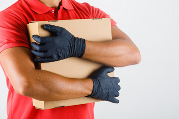 Delivery man in red uniform, medical gloves hugging cardboard box and looking confident , front view.