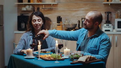 Relaxed young husband talking with his wife and pouring red wine in glasses. Romantic caucasian happy couple sitting at the table in kitchen celebrating marriage at candle lights, love and anniversary