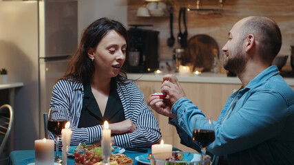 Handsome young man asking for marriage his girlfriend. Man making proposal to his bride fiancee to be in the kitchen during romantic dinner. Happy surprised woman smiling being speechless.
