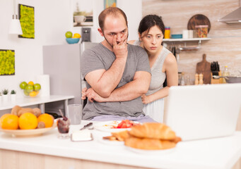 Obraz na płótnie Canvas Stressed entrepreneur working on laptop sitting on kitchen table while his wife is cooking breakfast. Unhappy, stressed, frustrated furious negative and upset freelancer in pajamas yelling during