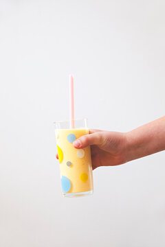 A child's hand holding a homemade mango lassi against a white wall