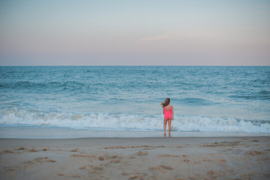 Small girl in a pink swimsuit faces the ocean at sunset.