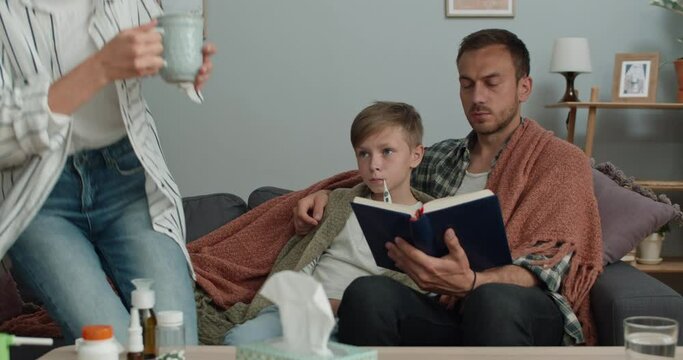 Dad Reading Book And His Sick Teen Son Measuring Temperature While They Sitting On Couch.Young Mom Taking Thermometer And Talking While Bringing Hot Drink . Concept Of Illness