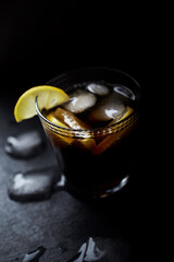 Glass of cola with lemon and ice cubes. Dark stone background. Selective focus. Close up.	