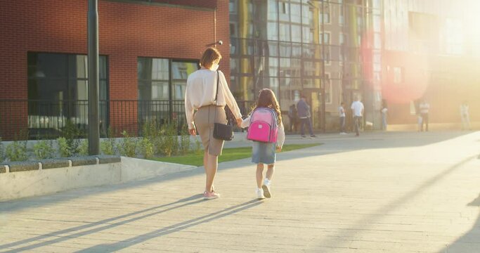 Rear of Caucasian beautiful mother and cute daughter in masks going to lessons outdoors in sun lights. Mom leads little junior student to school after quarantine. People on background. Pupil concept