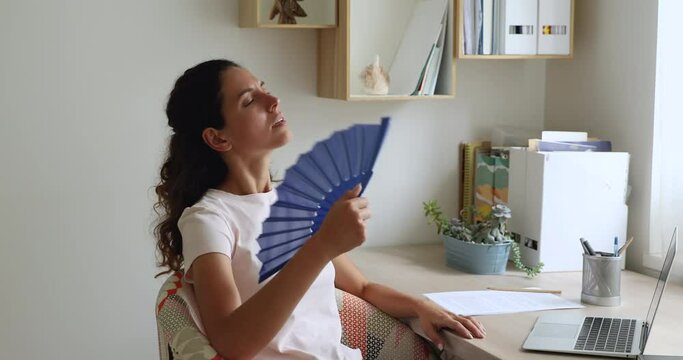 Woman sit at desk in front of laptop feels overheated due unbearable summer hot weather, holds waves handheld blue fan cooling herself. Apartment without air-conditioner, need climate control concept