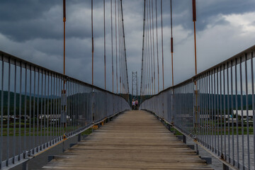 suspended pedestrian bridge with metal vertical cables and wooden path against the backdrop of village of Shamanka among Siberian mountains, cloudy landscape, geometric, symmetrical view