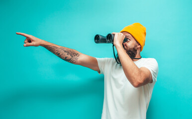 Young bearded hipster guy with binoculars over isolated blue wall wearing white blank t-shirt. Crazy emotions