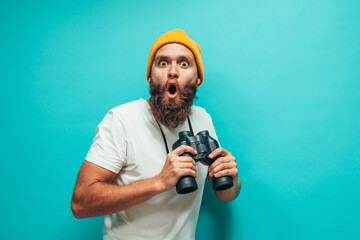 Young bearded hipster guy with binoculars over isolated blue wall wearing white blank t-shirt....