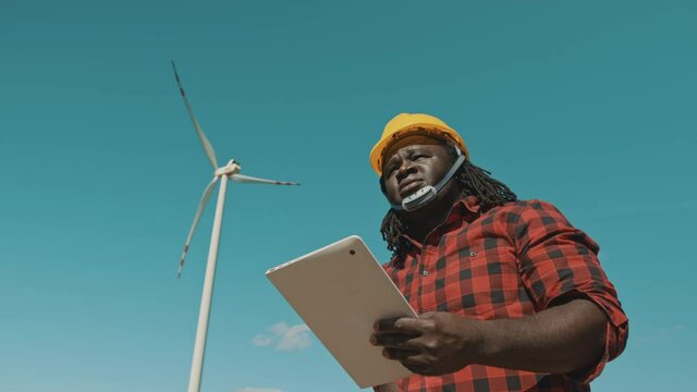 Professional african man in hard hat standing with modern digital tablet against windmill power station making inspections of objects. High quality 4k footage