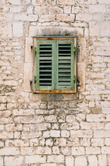 A window with closed, old, green, wooden shutters in the brick wall of the house.
