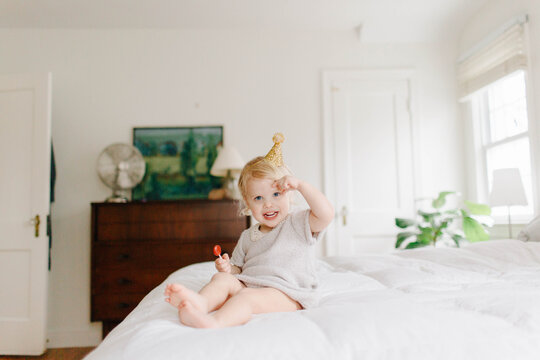 little girl sits on a bed wearing a party hat and having a lollipop to celebrate her birthday