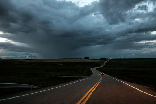 A long car drives toward a storm on a prairie road as dusk.