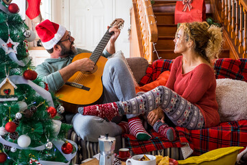 Happy young adult couple at home enjoy and celebrate christmas eve together having fun and love - people real life at home - man playing guitar for woman - husband and wife relationship with happiness