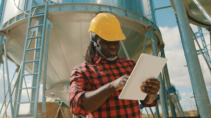Young handsome african man, engineer with safety helmet using tablet in front of silo system. High...