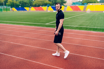 Fototapeta premium Handsome caucasian male in trendy wear and headphones walking on stadium listening favorite songs, portrait of hipster guy prepare for cardio workout enjoying online radio in sound accessory