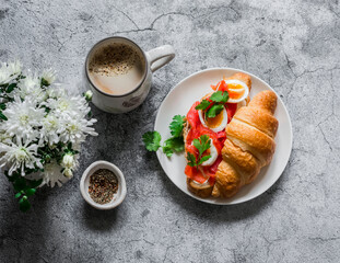 Delicious brunch - coffee with cream and croissant sandwich with cream cheese, smoked salmon and boiled egg on a grey background, top view