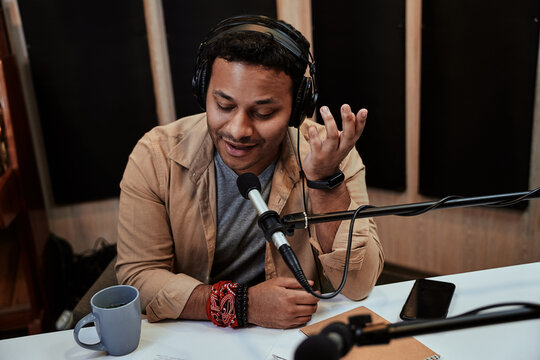 Happy young male radio host talking, broadcasting in studio using microphone and headphones
