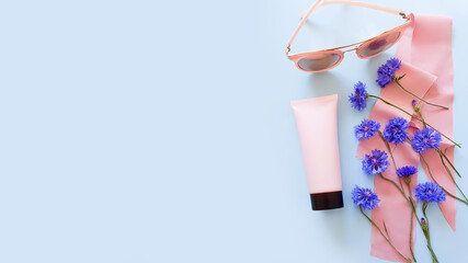 Mockup of pink squeeze bottle plastic tube with black cap, blue wild flowers, pink sunglasses and pink silk ribbon on a pastel blue background. Bottle for branding and label. Natural organic cosmetics