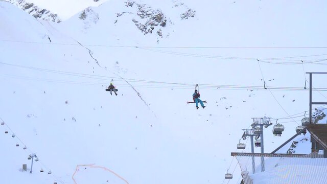 Snowboarders ride a zip line by aerial cableway, descend from the mountain to the base. Winter ski resort. Entertainment, recreation, vacation. Slow motion video