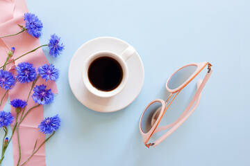 Top view of coffee cup, blue wild flowers, pink sunglasses and pink silk ribbon on a pastel blue background. Summer flatlay. Lifestyle, mock-up.