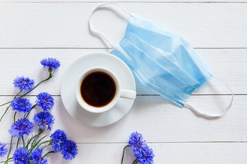 Medical protective blue face mask, coffee cup and blue flowers on white table. Biological concept of life in a new reality. Top view, flatlay lifestyle.