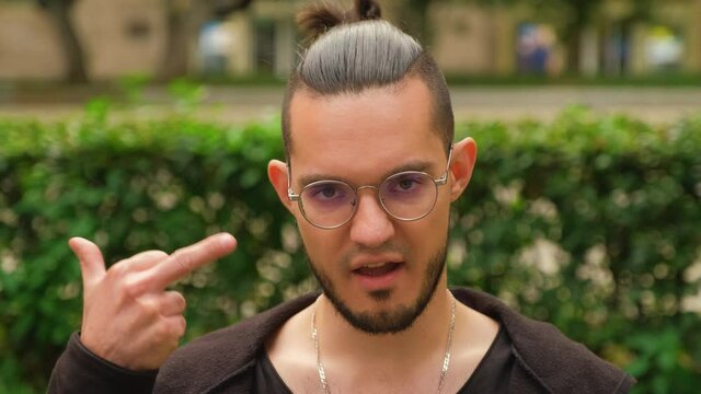 A charismatic dark-haired European with round glasses and light bristles chews gum insolently looking at the camera and makes an indecent gesture. Aggressive emotion.