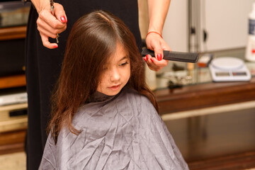 Fototapeta premium Portrait of a young girl getting her hair cut in home. Mom's hand with hairdressing scissors. Little child is getting a haircut in home during the lockdown.