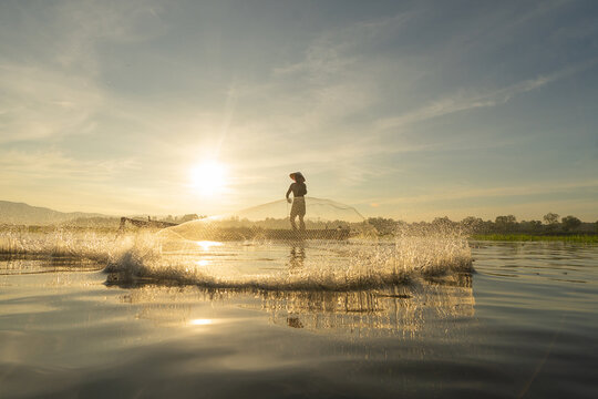 Silhouette Fisherman casting or throwing a net for catching freshwater fish in nature lake or river with reflection in morning time in Asia in Thailand. People.