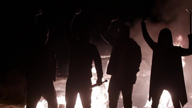 Silhouette of protesting guys with baseball bats and crowbar, raising their hands and shouting slogans, against background of smashed burning car