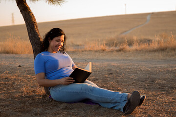 Young Caucasian woman reading a black book sitting on the ground in a park with sunset in the background