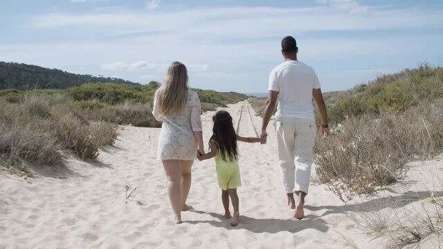 Parents and kid wearing summer clothes, walking on sand path, girl holding parents hands. Rear view. Family outdoors concept
