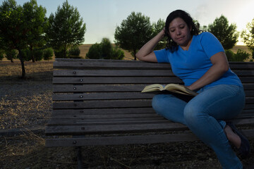 Obraz premium Caucasian woman sitting on a bench in the park reading a black book while wearing jeans and a blue blouse