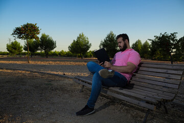 Caucasian man reading a book in the park while wearing a pink shirt and jeans