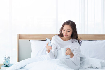 Morning girl with cup of coffee in hands in bed on a white blanket, simple, home, while playing with smartphone. Beautiful Asia woman drinking a coffee in her bed while reading from smartphone. 