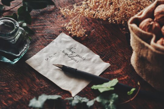 White peace of paper with "New Year's Resolutions" written on it. Yule winter solstice (Christmas) theme with nature themed background of dried flowers ivy branch, walnuts, ink bottle, fountain pen 