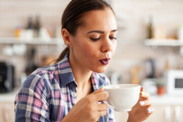Caucasian young woman trying to drink hot green tea. Close up of pretty lady sitting in the kitchen in the morning during breakfast time relaxing with tasty natural herbal tea from white teacup.
