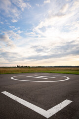 Fototapeta premium View of the private helipad on a warm summer evening. An asphalt helipad against the backdrop of a green field and a cloudy evening sky.
