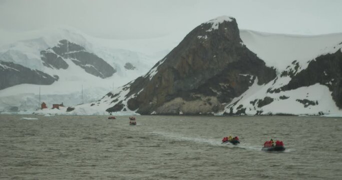 5 zodiacs (small boats) travel from the science base to the boat in Antarctica. A giant mountain and glacier can be seen in the background.
