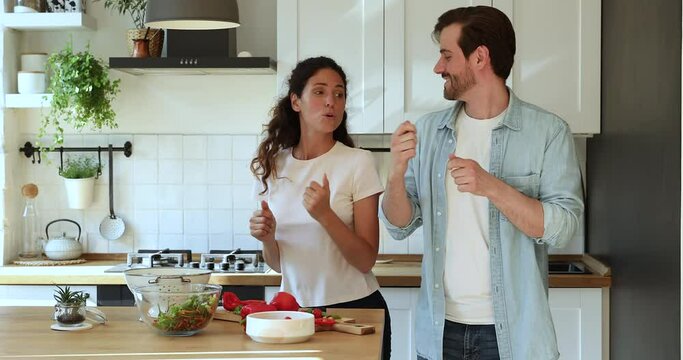Excited millennial 35s couple in love active wife and husband listening music dancing moving lively in domestic kitchen distracted from vegetarian food preparation. Healthy lifestyle and fun concept