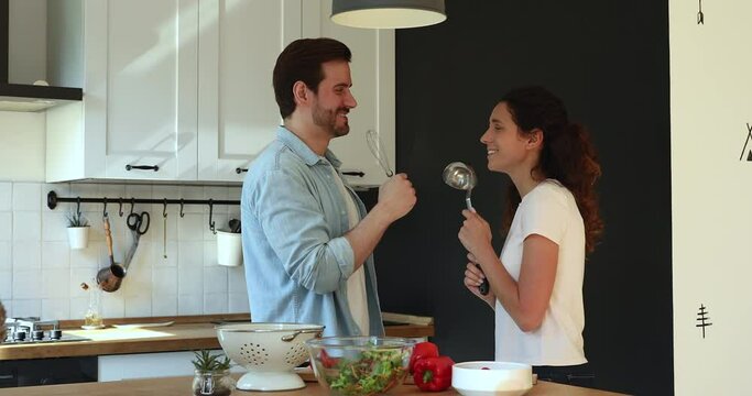 Happy couple cooking in kitchen dancing and singing karaoke using kitchenware like microphones enjoy food preparation together during romantic date at new home. Have fun, hobby and lifestyle concept
