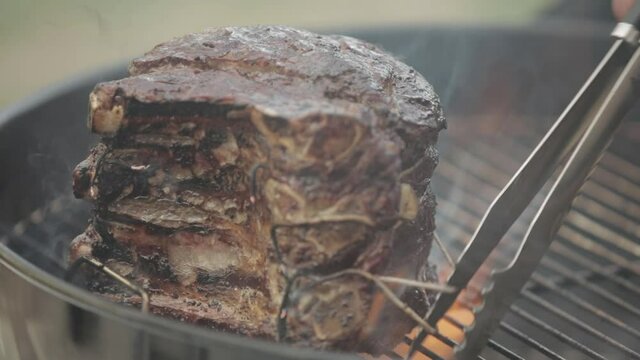 Tongs moving prime rib on grill as it cooks with smoke and flames and sizzles