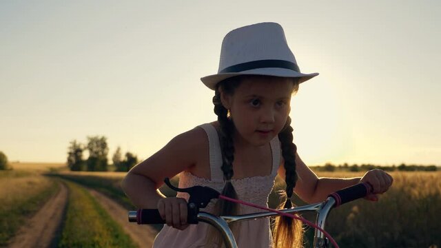 A girl in a cowboy hat rides a bicycle on the road in a field. The kid is enjoying the meadow landscape at sunset. The baby rides a bike and laughs. The child is dreaming.